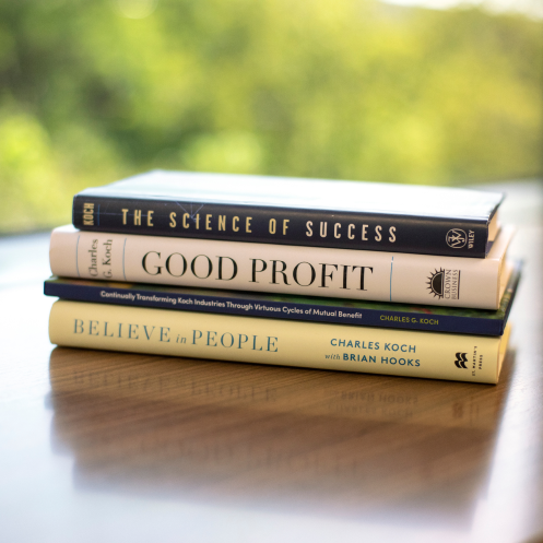 A neatly stacked pile of four books authored by Charles Koch on a wooden desk, with a blurred outdoor background.