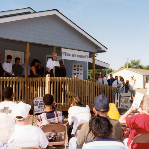 An archive photo of Charles Koch speaking from a podium addressing a crowd at a Koch Habitat for Humanity build site.